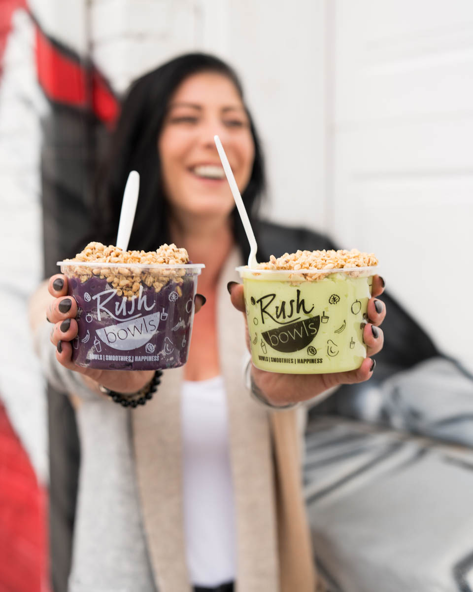 A Rush Bowls customer happily holding up acai smoothie bowls topped with granola.